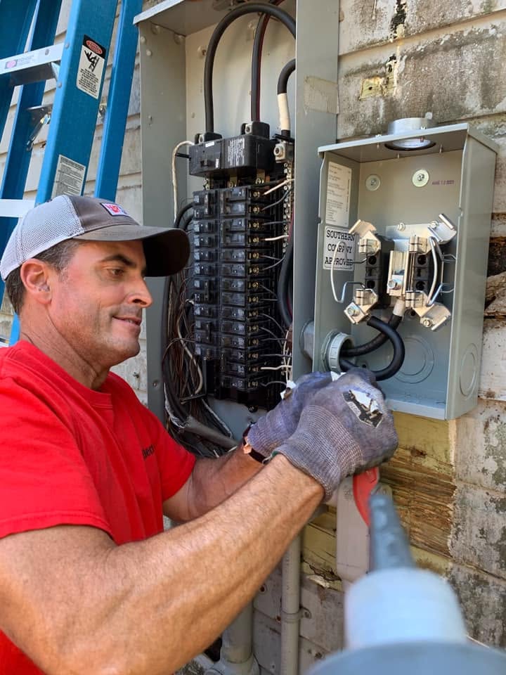 Jacob Brewster Master Electrician in a red shirt and gloves repairs open electrical panels on an outdoor wall, focused amid exposed wiring and tools.