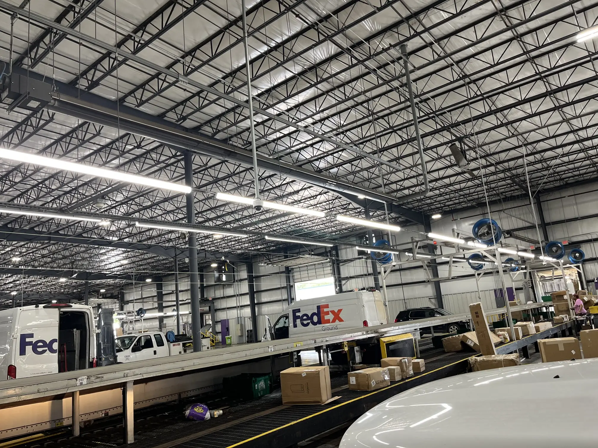 jb electric mobile al installing lighting Inside a busy FedEx distribution warehouse, conveyor belts carry cardboard packages beneath bright lights and a steel-beam ceiling.