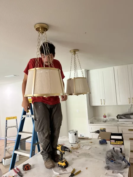 Worker installs a brass-and-beige pendant light in a kitchen renovation, standing on a ladder amid tools, cabinets, and construction debris.
